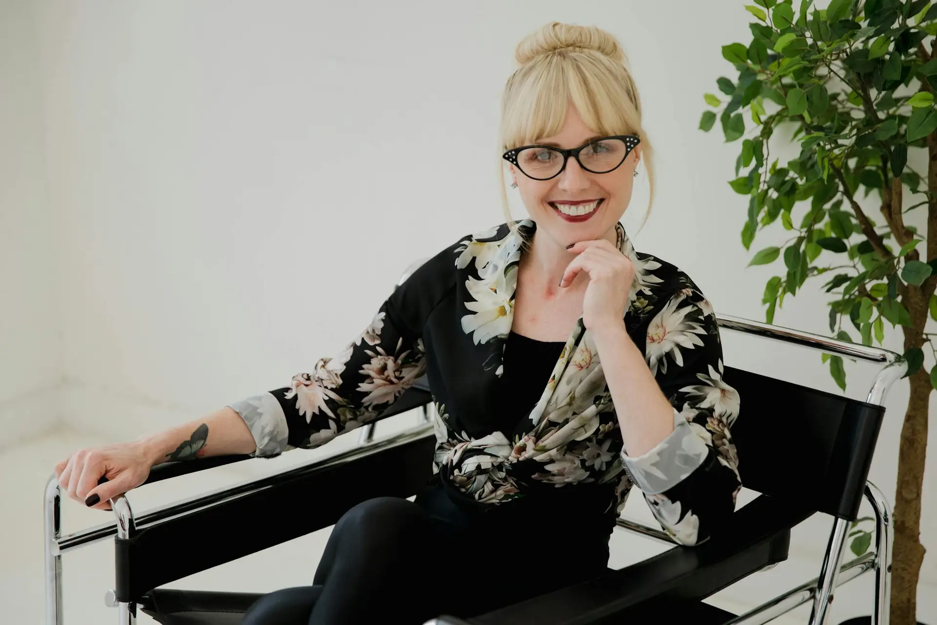 Blonde woman with eyewear smiling and sitting with hand on chin in a floral shirt, indoors.