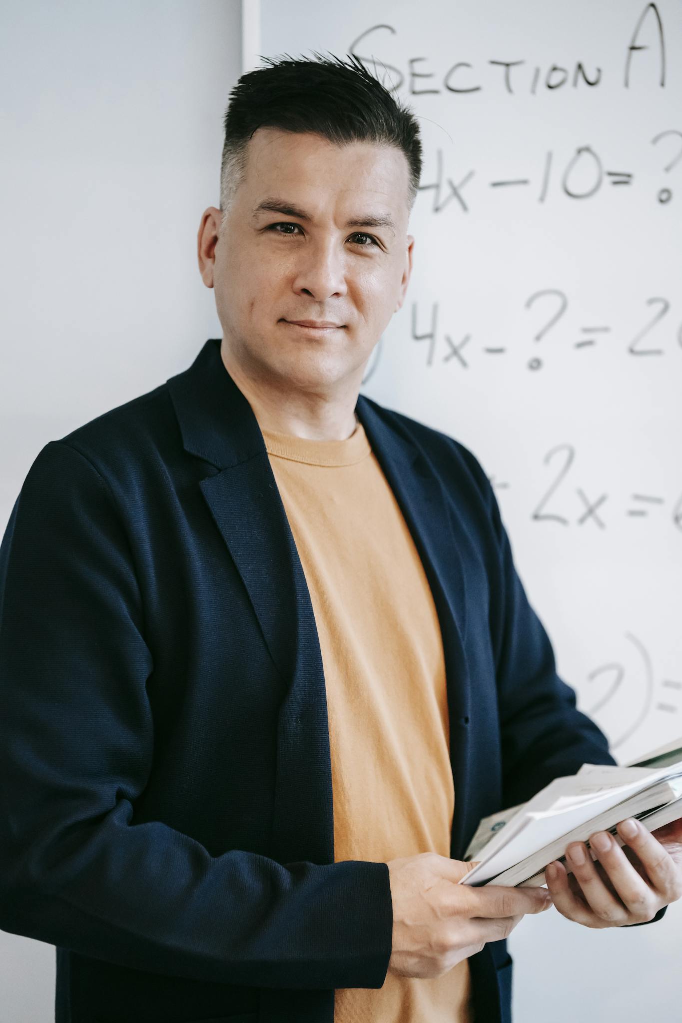 Male teacher standing in a classroom, holding books with a smile in front of a whiteboard.