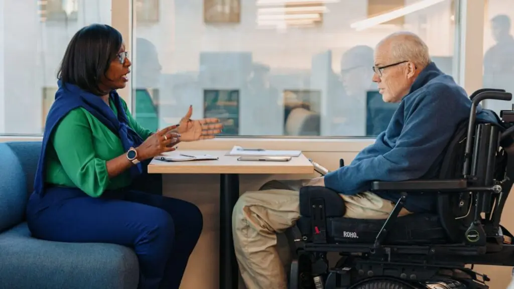 Woman and older man using a wheelchair talk at a table by a window.