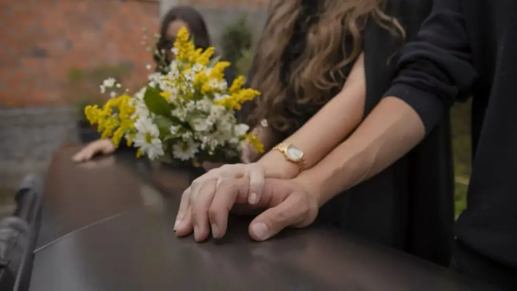 Two people rest their hands on a casket with a bouquet of yellow and white flowers.