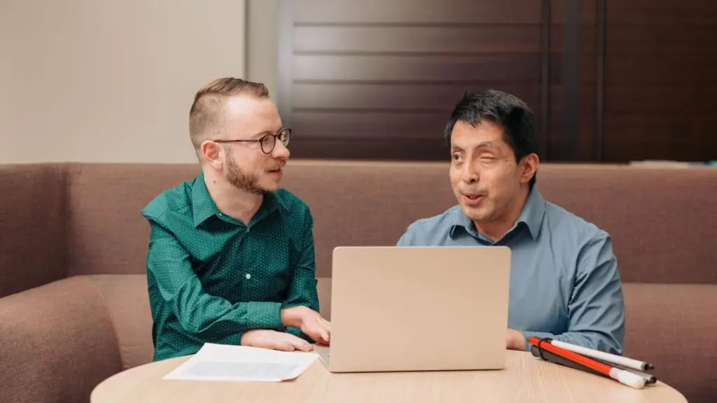 Two people at a table talking and working together on a laptop.