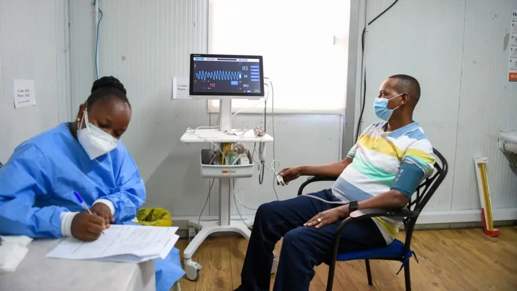 Healthcare worker takes notes while a masked patient sits beside a vital-signs monitor.