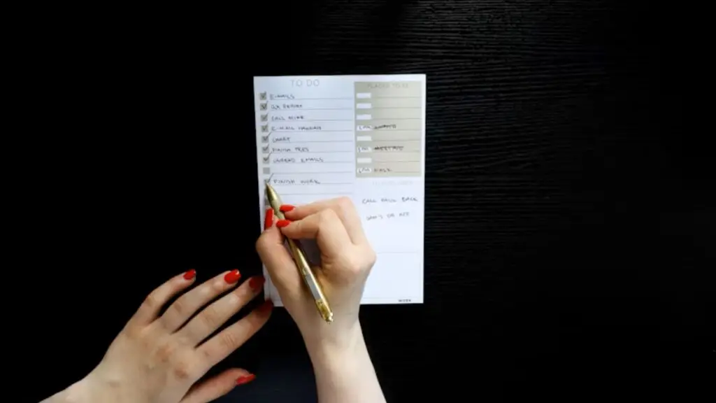 Person writing on a to-do list with a gold pen on a dark desk.